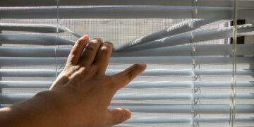 A hand gently opens window blinds, letting sunlight stream indoors.