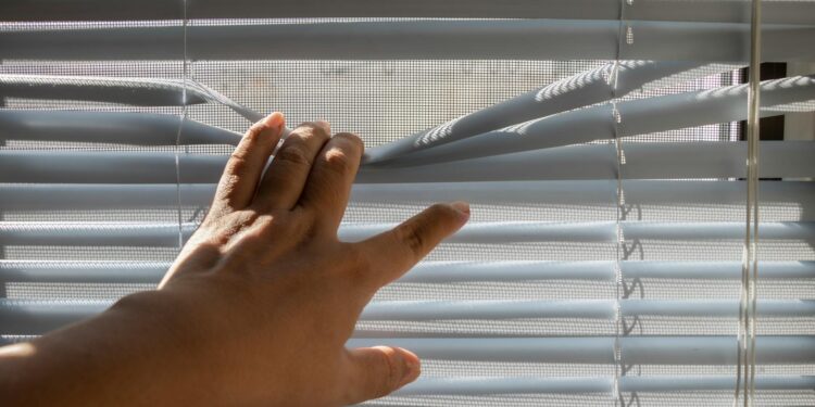 A hand gently opens window blinds, letting sunlight stream indoors.