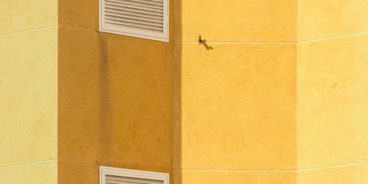 Close-up of a modern yellow building corner featuring ventilation grilles for architectural interest.