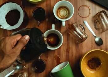Hand pouring coffee into various cups with spills on wooden table.