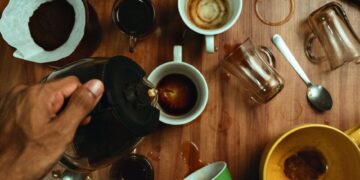Hand pouring coffee into various cups with spills on wooden table.