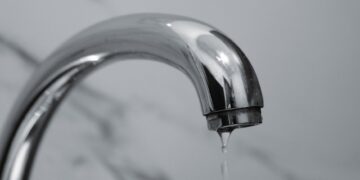 A close-up shot of a modern chrome bathroom faucet with water dripping from the tip.