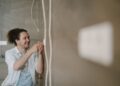An electrician with curly hair adjusts an outlet indoors, smiling as he works.