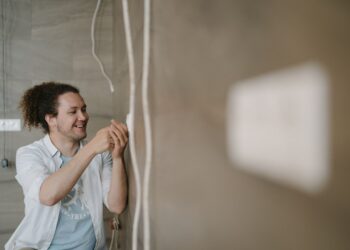 An electrician with curly hair adjusts an outlet indoors, smiling as he works.