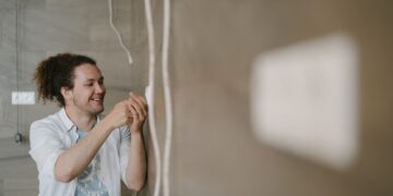 An electrician with curly hair adjusts an outlet indoors, smiling as he works.