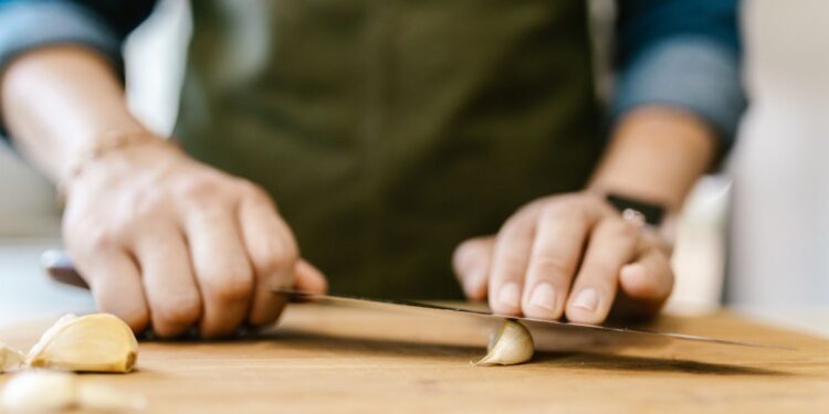 Close-up of a person slicing garlic on a wooden cutting board, emphasizing cooking preparation.
