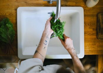Tattooed hands washing fresh herbs in a kitchen sink, highlighting daily culinary routines.