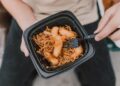 Tasty Asian noodles with shrimp in a black container held by a person.