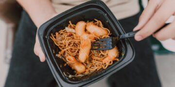 Tasty Asian noodles with shrimp in a black container held by a person.