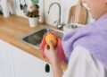 Crop unrecognizable person in casual clothes standing and wiping ripe fresh lemon with cloth in light kitchen in daytime