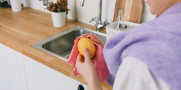 Crop unrecognizable person in casual clothes standing and wiping ripe fresh lemon with cloth in light kitchen in daytime