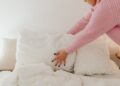 A woman in a pink sweater adjusts white pillows in a bright bedroom.
