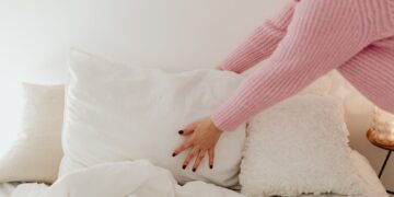 A woman in a pink sweater adjusts white pillows in a bright bedroom.