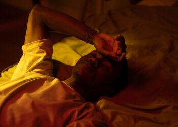 A man lying in bed at night with a moody expression and warm lighting.