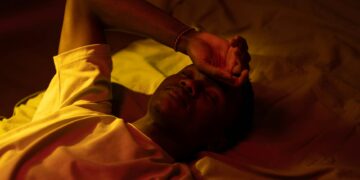 A man lying in bed at night with a moody expression and warm lighting.