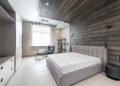 Bed with white mattress placed at wall with decorative element against cupboards in light stylish bedroom with window and armchair