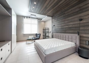 Bed with white mattress placed at wall with decorative element against cupboards in light stylish bedroom with window and armchair