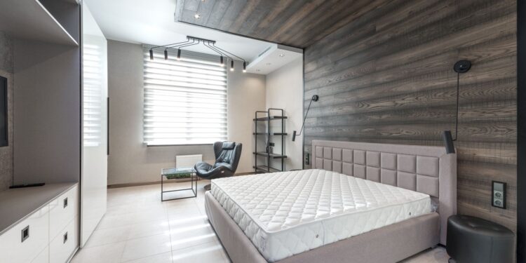 Bed with white mattress placed at wall with decorative element against cupboards in light stylish bedroom with window and armchair