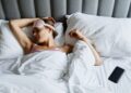 Woman resting in bed wearing an eye mask with a smartphone nearby, embracing relaxation.