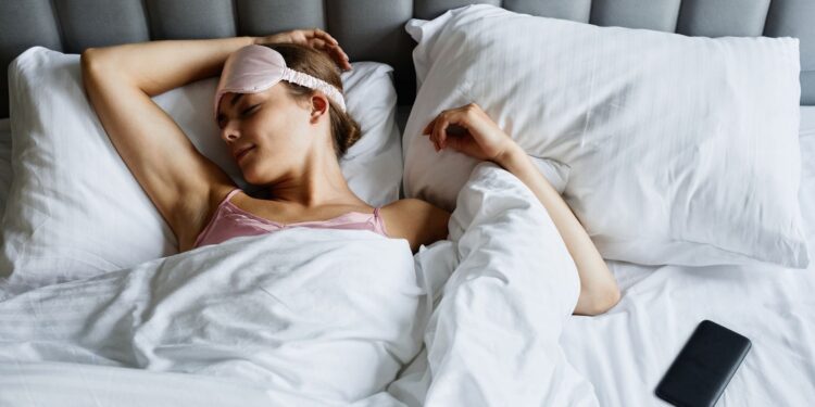 Woman resting in bed wearing an eye mask with a smartphone nearby, embracing relaxation.
