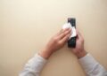 Hands cleaning a TV remote control with a cloth on a beige background for hygiene and cleanliness.