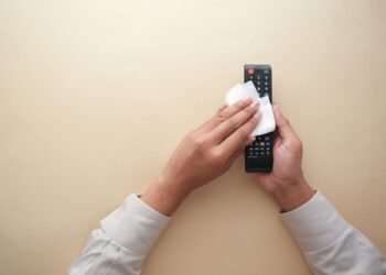 Hands cleaning a TV remote control with a cloth on a beige background for hygiene and cleanliness.