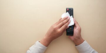 Hands cleaning a TV remote control with a cloth on a beige background for hygiene and cleanliness.