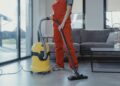 A professional cleaner in red coveralls vacuuming a modern living room with a yellow vacuum cleaner.