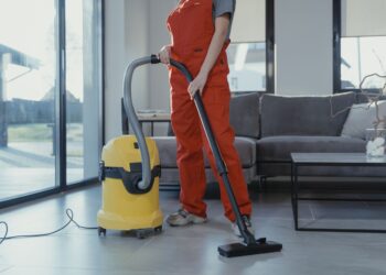 A professional cleaner in red coveralls vacuuming a modern living room with a yellow vacuum cleaner.