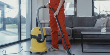 A professional cleaner in red coveralls vacuuming a modern living room with a yellow vacuum cleaner.