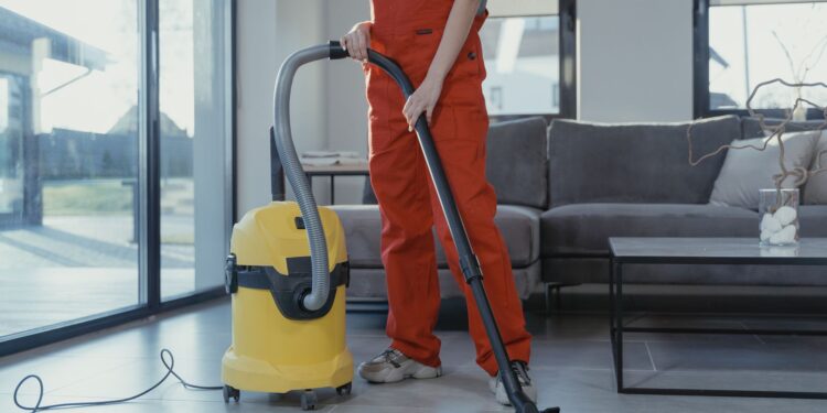 A professional cleaner in red coveralls vacuuming a modern living room with a yellow vacuum cleaner.