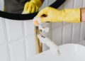 Anonymous person wearing yellow latex gloves washing golden faucet with sponge in modern bathroom while cleaning house