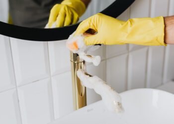 Anonymous person wearing yellow latex gloves washing golden faucet with sponge in modern bathroom while cleaning house