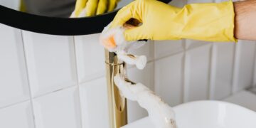 Anonymous person wearing yellow latex gloves washing golden faucet with sponge in modern bathroom while cleaning house
