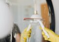 Anonymous person in yellow rubber gloves wiping water by squeegee from mirror in bathroom