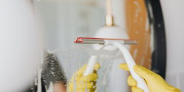 Anonymous person in yellow rubber gloves wiping water by squeegee from mirror in bathroom