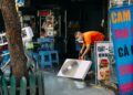 A man cleans a fan on a vibrant urban street with market stalls.