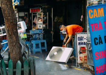 A man cleans a fan on a vibrant urban street with market stalls.