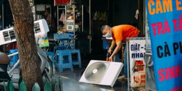 A man cleans a fan on a vibrant urban street with market stalls.