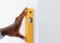 A close-up of a hand using a yellow level tool against a white wall to ensure vertical alignment.