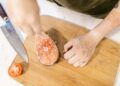 A person preparing salmon steak on a wooden board with a knife and tomato slice.
