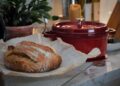Close-up of a rustic loaf of bread on a kitchen counter with a red Dutch oven nearby.