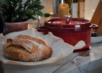 Close-up of a rustic loaf of bread on a kitchen counter with a red Dutch oven nearby.