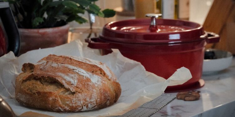 Close-up of a rustic loaf of bread on a kitchen counter with a red Dutch oven nearby.