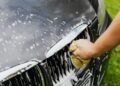 Detailed view of a hand washing a car grille with a soapy sponge outdoors.