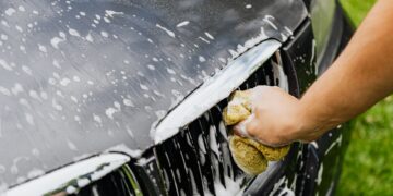 Detailed view of a hand washing a car grille with a soapy sponge outdoors.