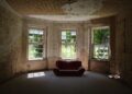 A decayed room featuring a vintage sofa and old windows letting in sunlight.
