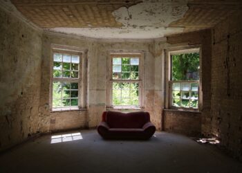 A decayed room featuring a vintage sofa and old windows letting in sunlight.