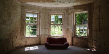 A decayed room featuring a vintage sofa and old windows letting in sunlight.
