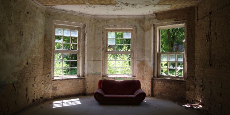 A decayed room featuring a vintage sofa and old windows letting in sunlight.